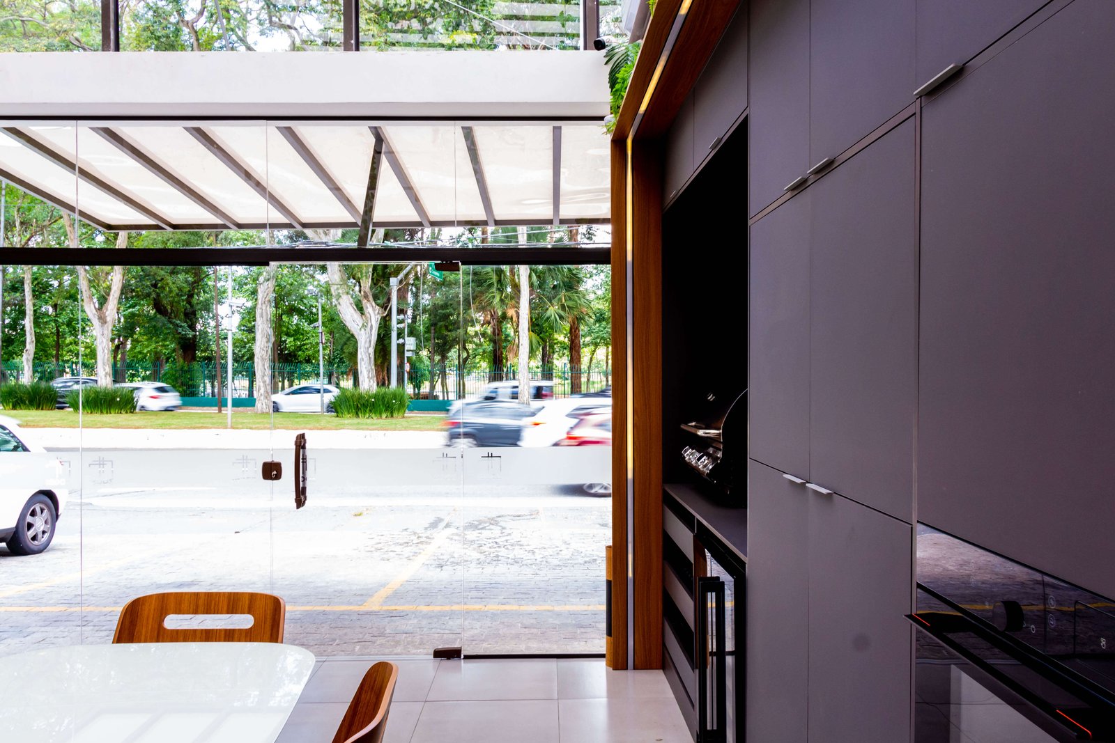 Perspectiva de dentro para fora do showroom, olhando através da fachada de vidro para a rua arborizada. A iluminação natural inunda o ambiente, iluminando os móveis cinza chumbo à direita.