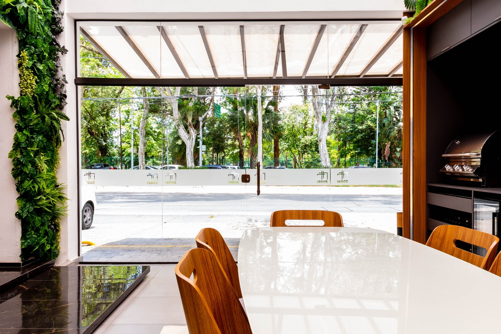 Vista interna do showroom olhando para a rua através de grandes portas de vidro com a logomarca jateada. Em primeiro plano, parte de uma mesa branca com cadeiras de madeira.