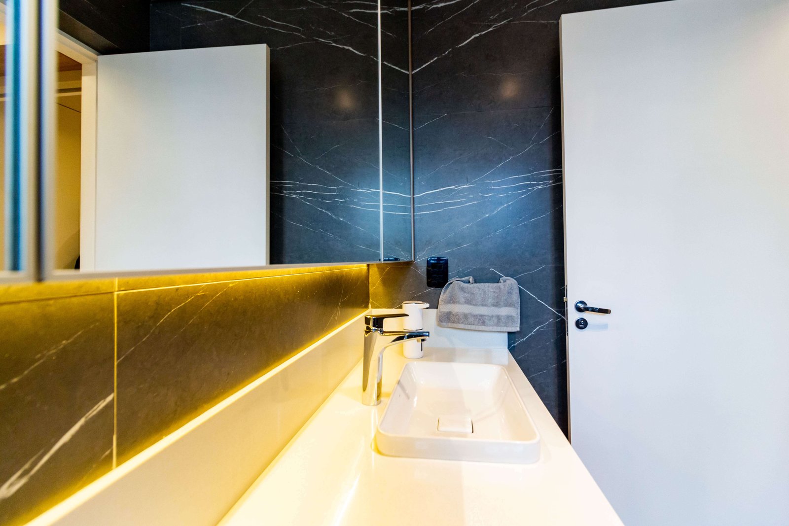 Lavabo luxuoso com revestimento de parede em mármore preto com veios brancos (Nero Marquina). O espelho possui iluminação indireta quente (backlight) e a torneira é cromada sobre uma cuba retangular branca.