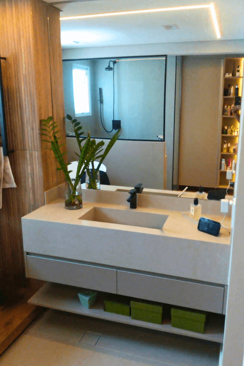 Bancada de banheiro em pedra bege com cuba esculpida e torneira preta moderna. O espelho reflete uma parede com revestimento ripado de madeira e o box ao fundo.