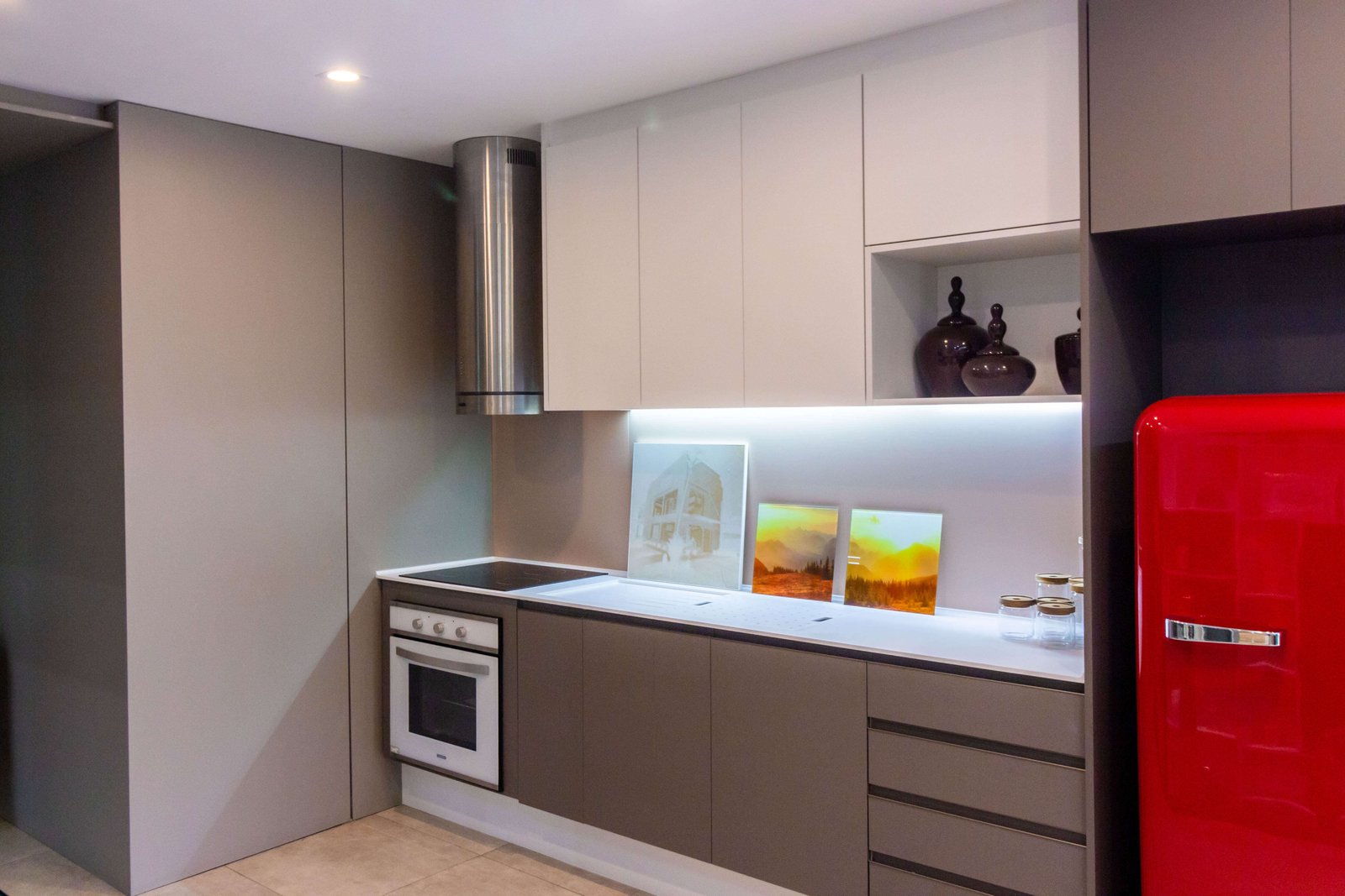 Uma parede de cozinha moderna com armários inferiores em tom de cinza (fendi) e superiores brancos. Vê-se um forno embutido, cooktop e uma coifa cilíndrica em inox.