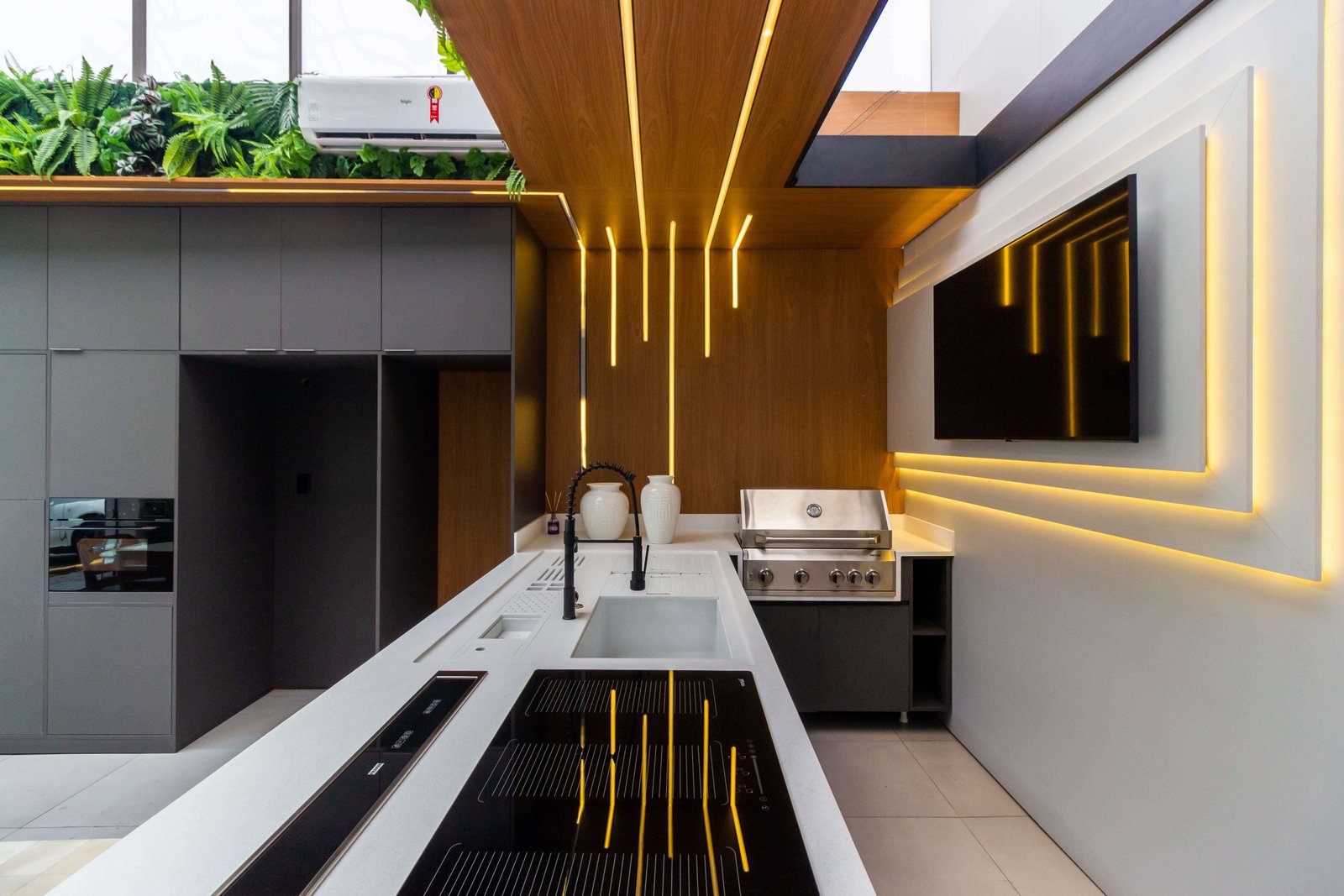 Foto de uma área gourmet luxuosa e moderna com teto alto. Em destaque, um painel de madeira que desce do teto para a parede com fitas de LED embutidas criando linhas verticais de luz. No centro, uma ilha branca com cooktop preto e torneira gourmet. À esquerda, armários planejados cinza escuro com forno embutido e plantas no topo. À direita, churrasqueira a gás em inox e TV montada em painel iluminado.
