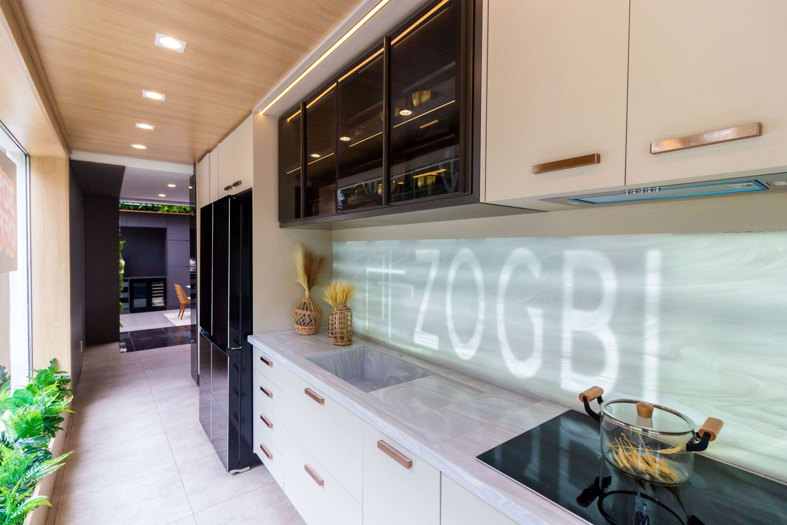 Uma cozinha de showroom com armários inferiores brancos e puxadores em tom cobre/rosé. Os armários superiores têm portas de vidro reflexivo escuro (bronze) e o frontão da pia é iluminado, exibindo a marca 'ZOGBI'.