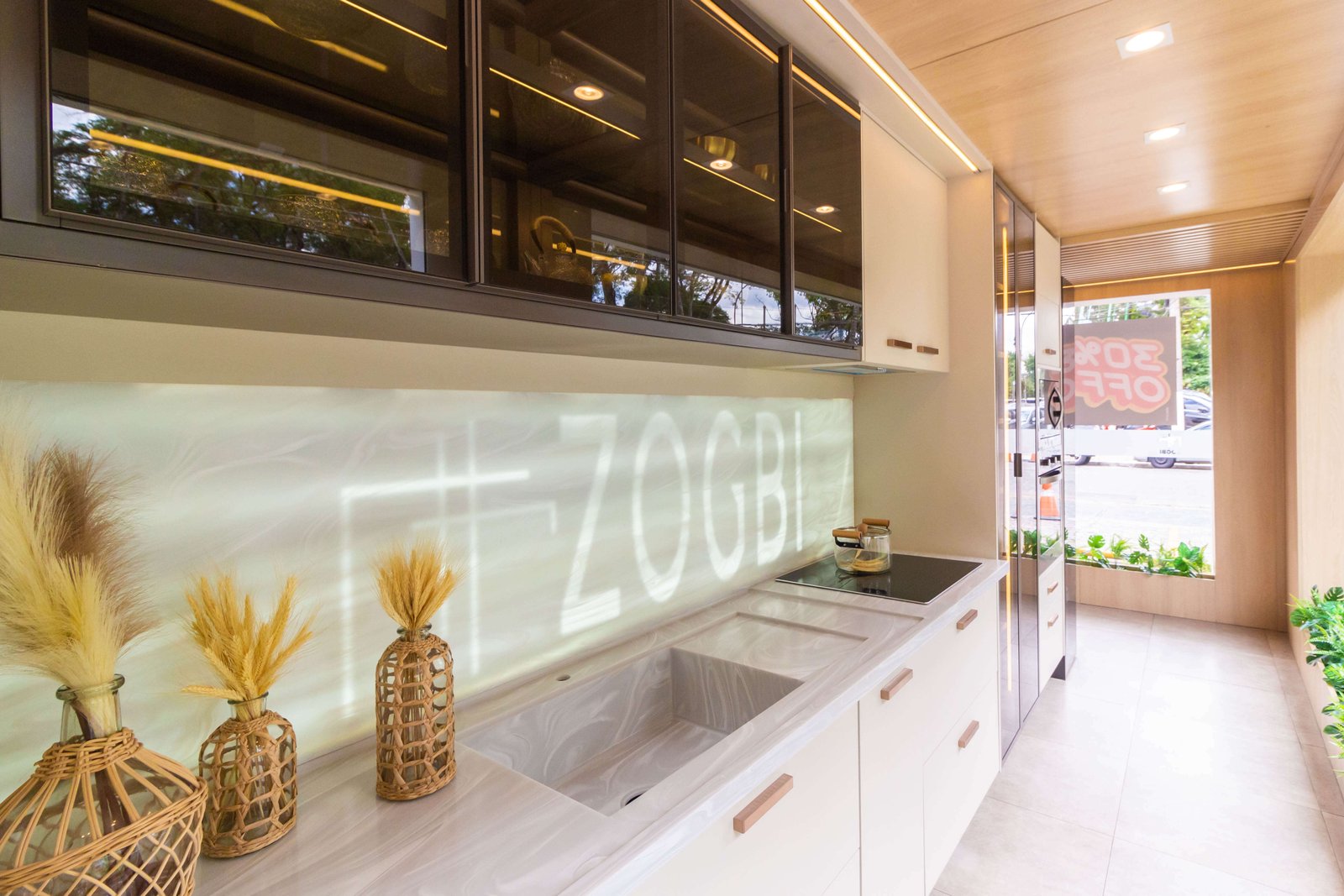Vista mais ampla da cozinha do showroom, mostrando a cuba esculpida na pedra, a cristaleira lateral com portas de vidro e iluminação interna, e novamente o destaque para a marca Zogbi no painel iluminado.