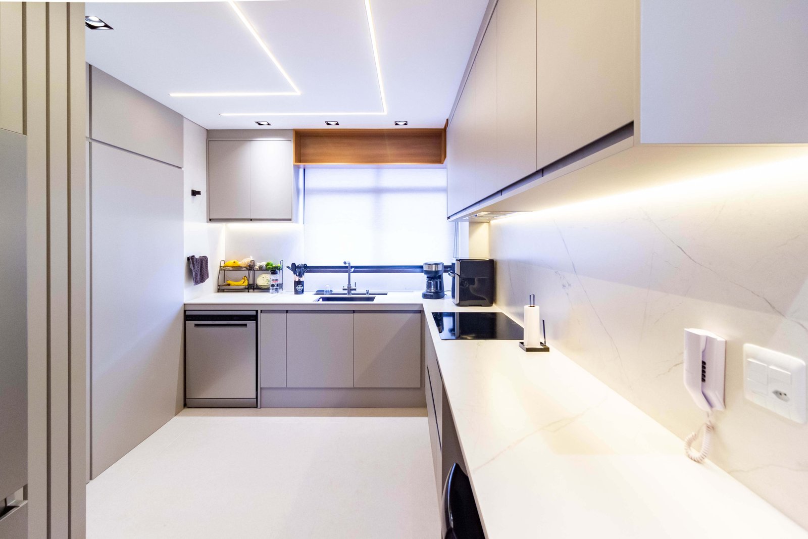 Vista geral da cozinha em formato de 'U', mostrando a bancada de pedra branca, os armários inferiores cinzas e a ampla janela ao fundo. O teto destaca-se pelo design de iluminação com perfis de LED quadrados.