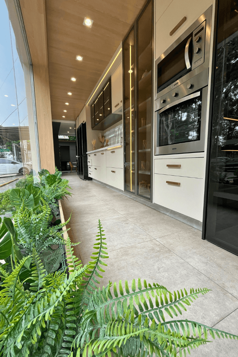 Uma vista em perspectiva de um corredor de cozinha (provavelmente showroom), com piso de porcelanato cinza e plantas à esquerda. À direita, vê-se uma torre de fornos, uma grande cristaleira iluminada com portas de vidro do chão ao teto e a bancada de pia ao fundo.