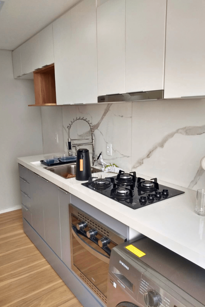 Bancada de trabalho com pedra branca e frontão (backsplash) alto em porcelanato marmorizado com veios cinzas. Vê-se uma torneira gourmet flexível, cuba de inox e cooktop a gás.