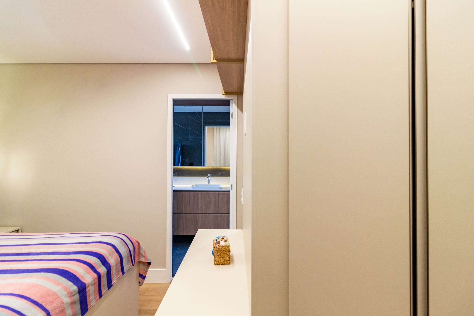 Vista da entrada de uma suíte, olhando para o banheiro ao fundo. O corredor tem armários brancos minimalistas e, pela porta aberta, vê-se o gabinete do banheiro em madeira escura.