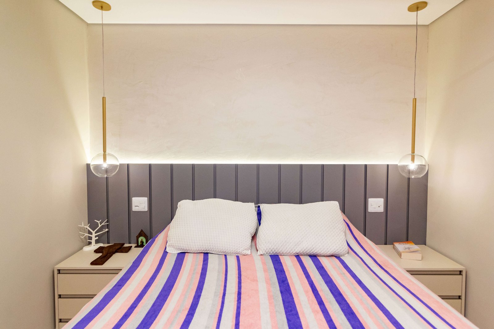Detalhe da cabeceira estofada cinza com gomos verticais, iluminada por fita de LED embutida. Um pendente moderno dourado desce sobre a mesa de cabeceira.