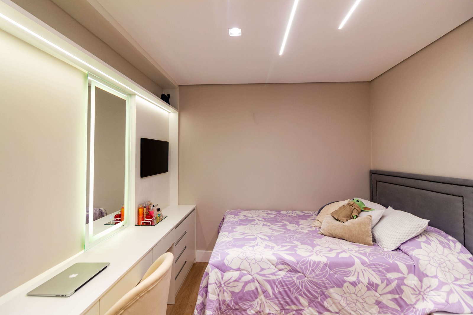Vista geral do quarto, mostrando a cama com colcha lilás estampada ao lado da bancada de estudos/beleza. O teto possui rasgos de luz lineares.