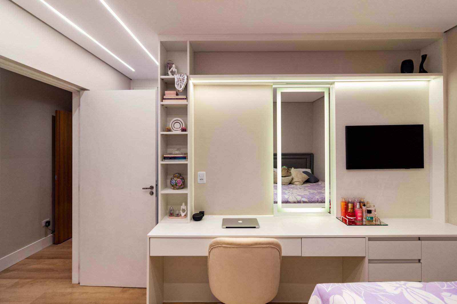 Vista frontal da estante e bancada branca. O móvel possui uma torre de nichos à esquerda, uma porta de correr com espelho iluminado no centro e espaço para TV à direita.
