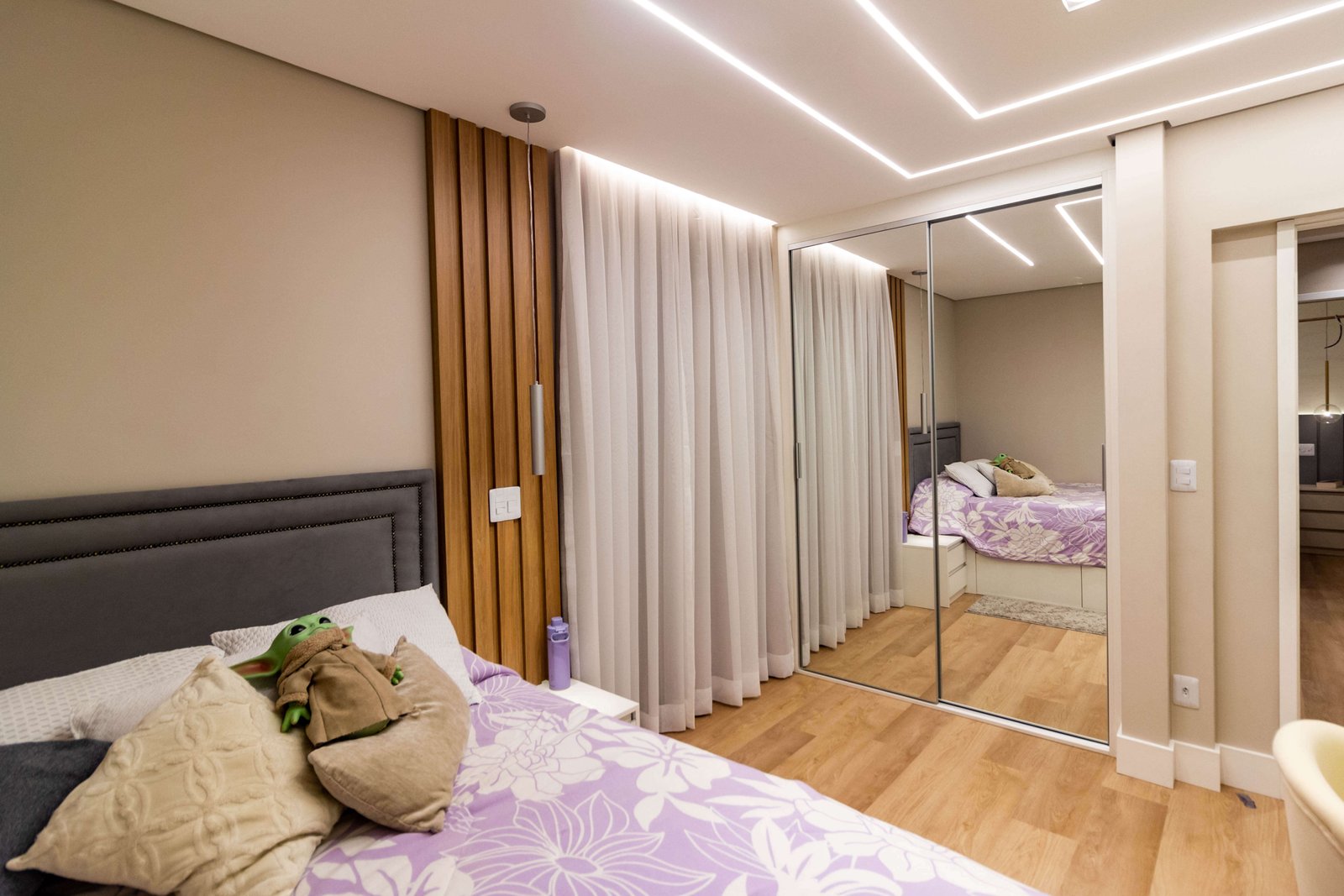 Um quarto juvenil aconchegante com roupa de cama lilás e uma pelúcia (Baby Yoda) sobre a cama. Ao lado, um guarda-roupa com portas de correr totalmente espelhadas reflete o painel ripado de madeira e a iluminação de LED no teto.