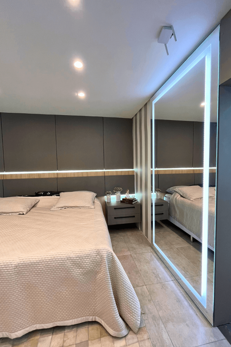 Um espelho de corpo inteiro com moldura de luz de LED intensa, posicionado ao lado da cama, criando profundidade e modernidade no quarto.