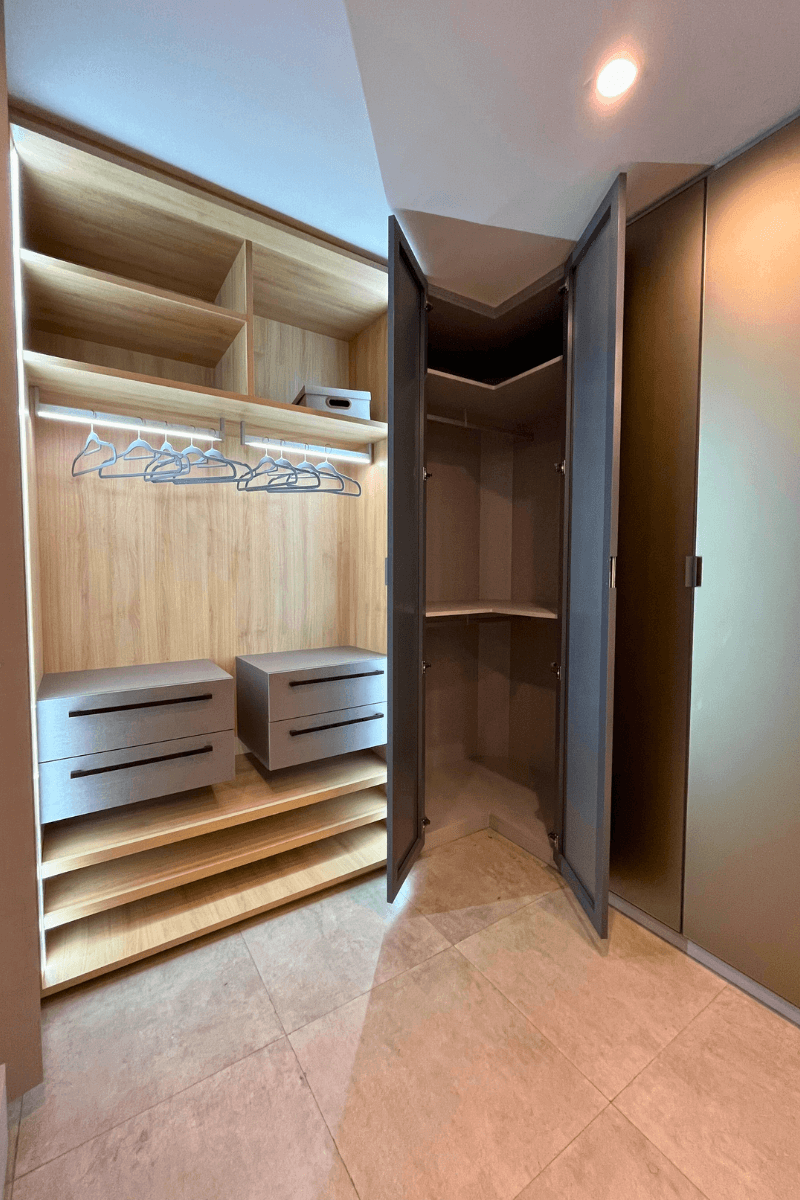 Um guarda-roupa de canto com as portas cinza-escuro abertas, revelando o interior em madeira clara. Há cabideiros iluminados por LED e gavetas suspensas, otimizando o ângulo do móvel.