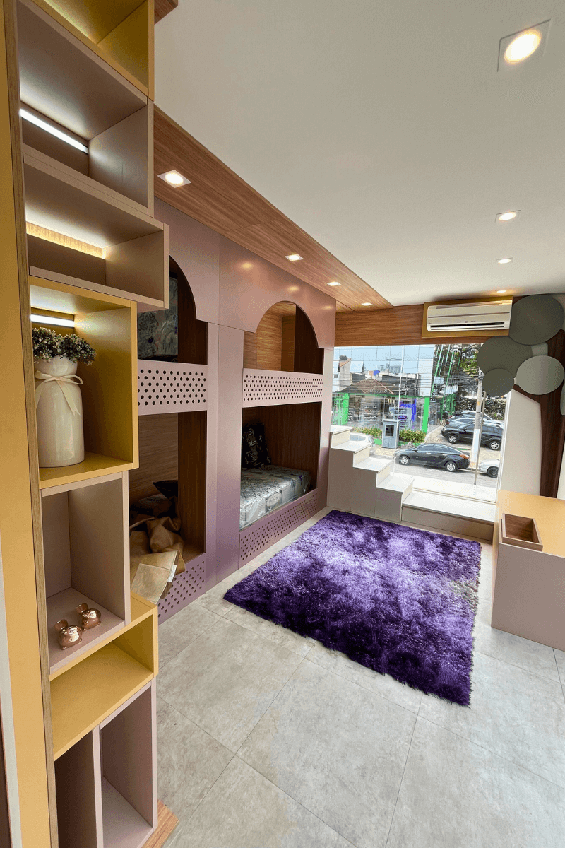 Um quarto infantil lúdico e colorido. Destaca-se uma beliche com estrutura de marcenaria rosa em formato de casinha/castelo, um tapete felpudo roxo vibrante e uma estante lateral com nichos amarelos.
