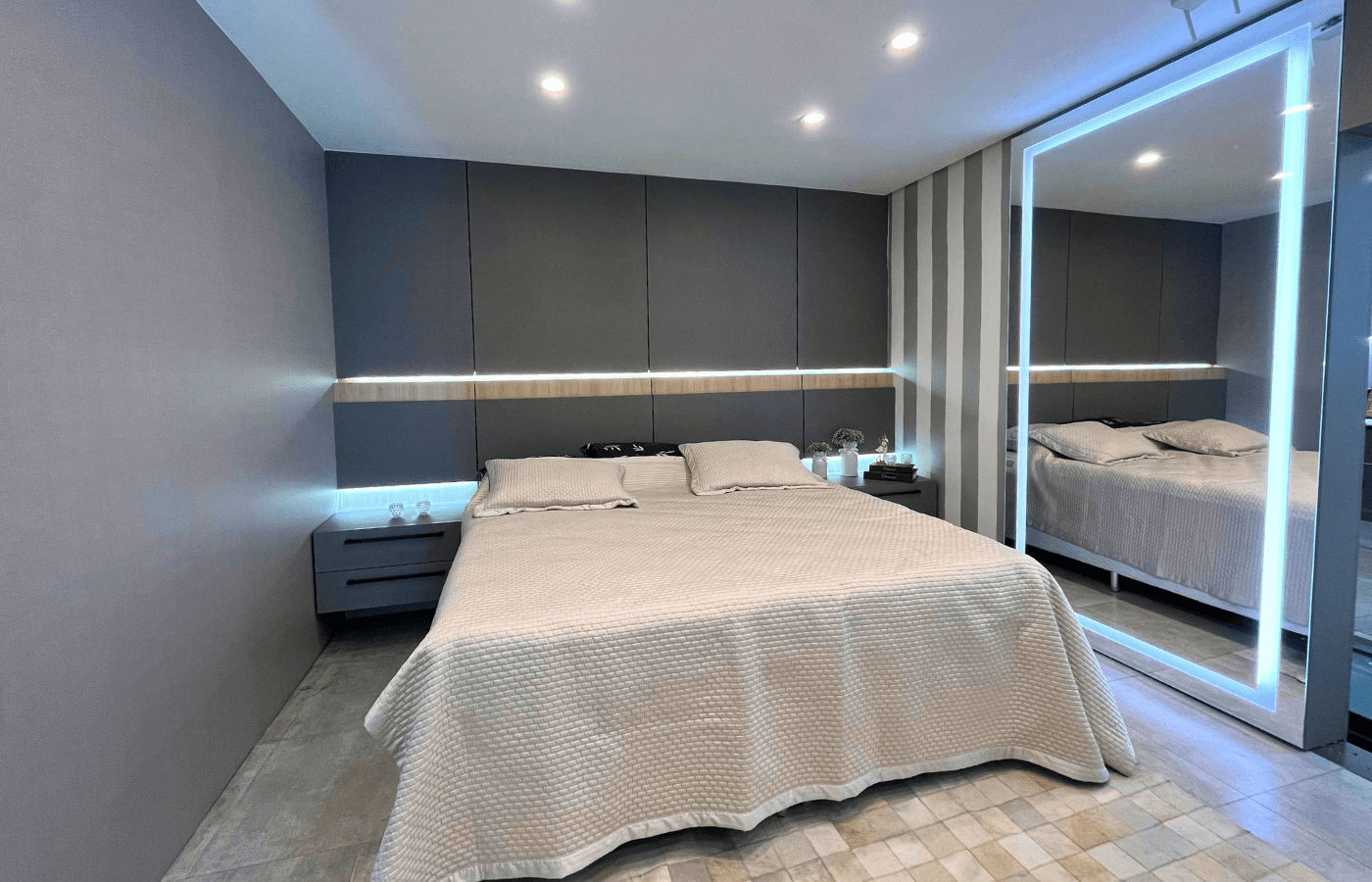 Outro ângulo do mesmo quarto de casal, mostrando a cama king size, o painel de cabeceira estofado que cobre toda a parede e um grande espelho de chão com moldura de LED.