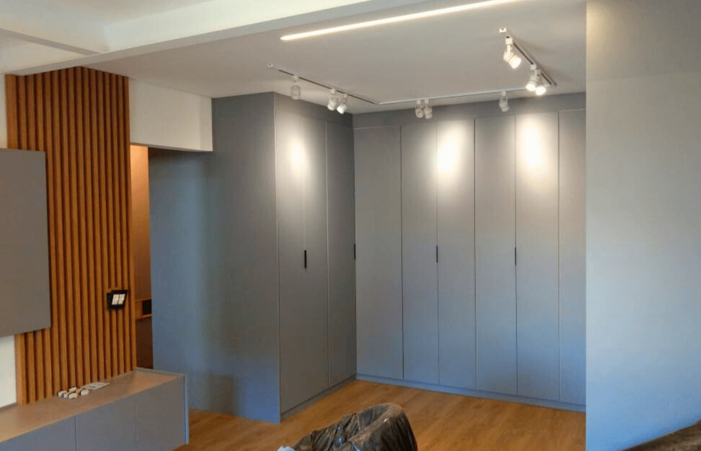 Um conjunto de armários de canto em cinza liso, cobrindo duas paredes inteiras (formato em L). A iluminação é feita por trilhos de spots no teto.