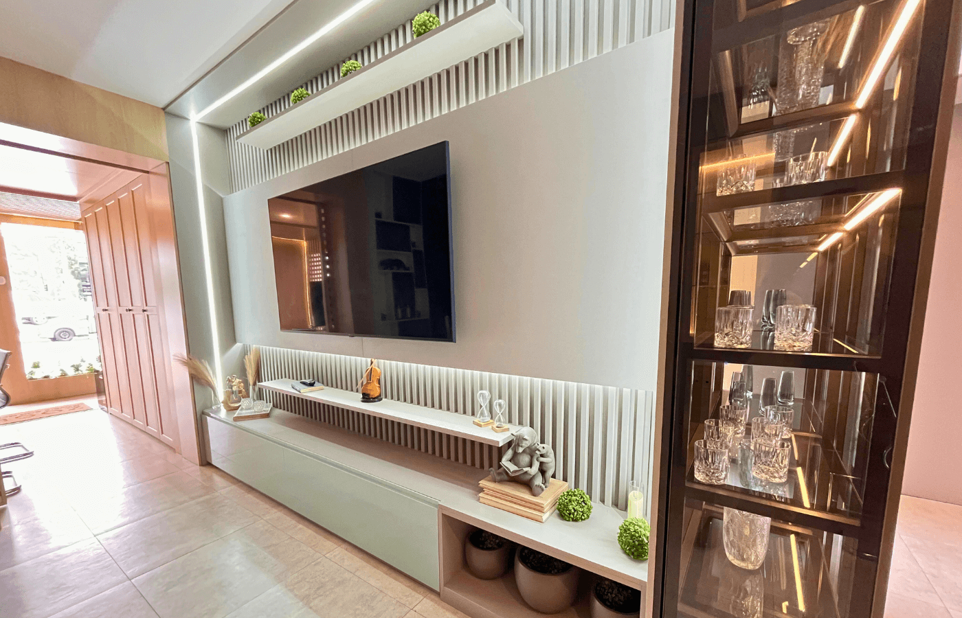 Vista lateral de um home theater luxuoso com painel ripado claro ao fundo, prateleiras iluminadas por LED e uma cristaleira lateral com portas de vidro bronze.