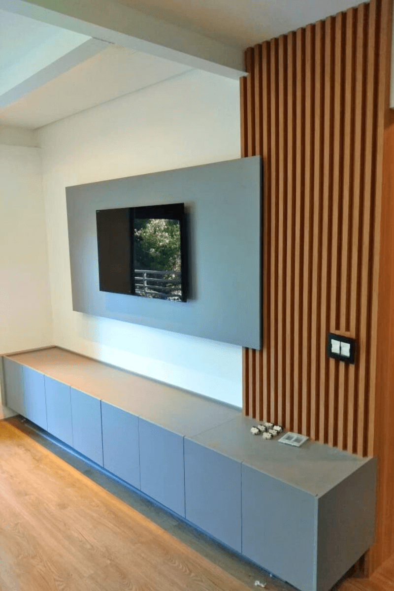 Um design minimalista composto por um painel cinza liso centralizado para a TV, um rack baixo e longo na mesma cor, e um detalhe vertical em madeira ripada na lateral direita.