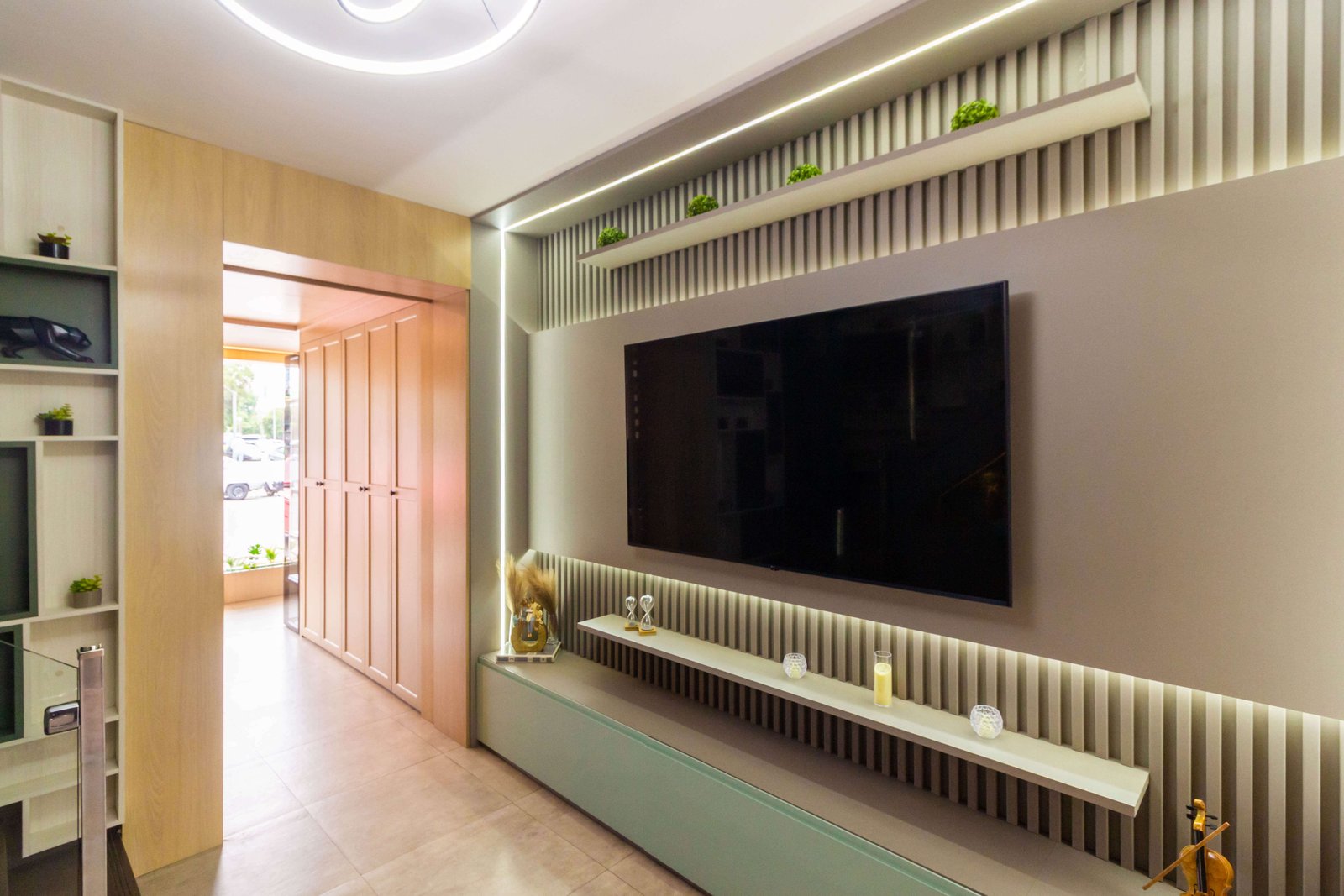 Vista frontal do primeiro ambiente, destacando o painel da TV cinza sobreposto ao fundo ripado, com iluminação de LED emoldurando a marcenaria e prateleira superior com plantas.
