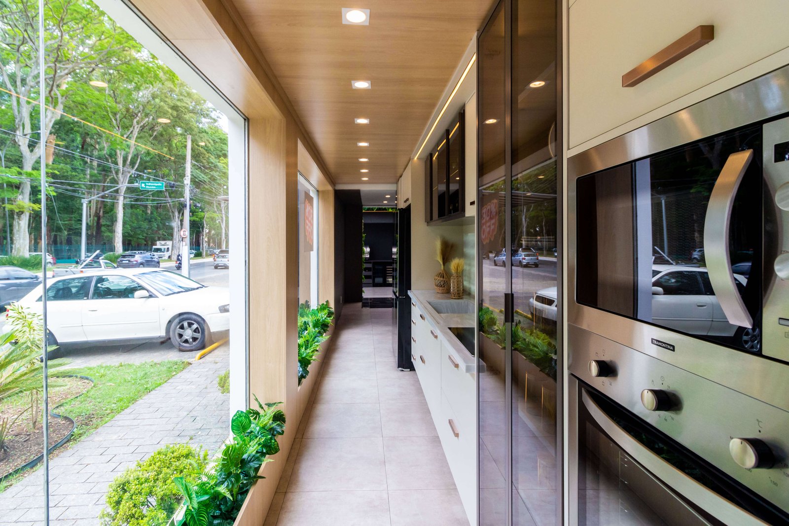Ambiente planejado em corredor com armários embutidos brancos, torre de fornos e teto rebaixado em madeira com iluminação, integrado a grandes janelas com vista para o verde.