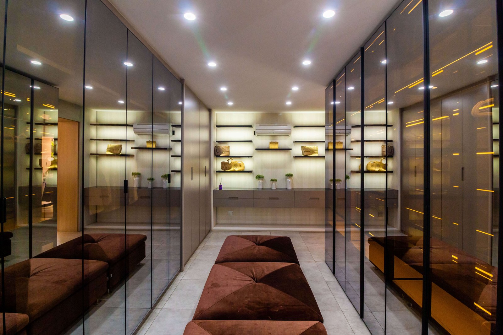 Closet planejado com portas de vidro fumê, iluminação interna em LED e ilha central acolchoada, demonstrando sofisticação e organização em móveis sob medida.