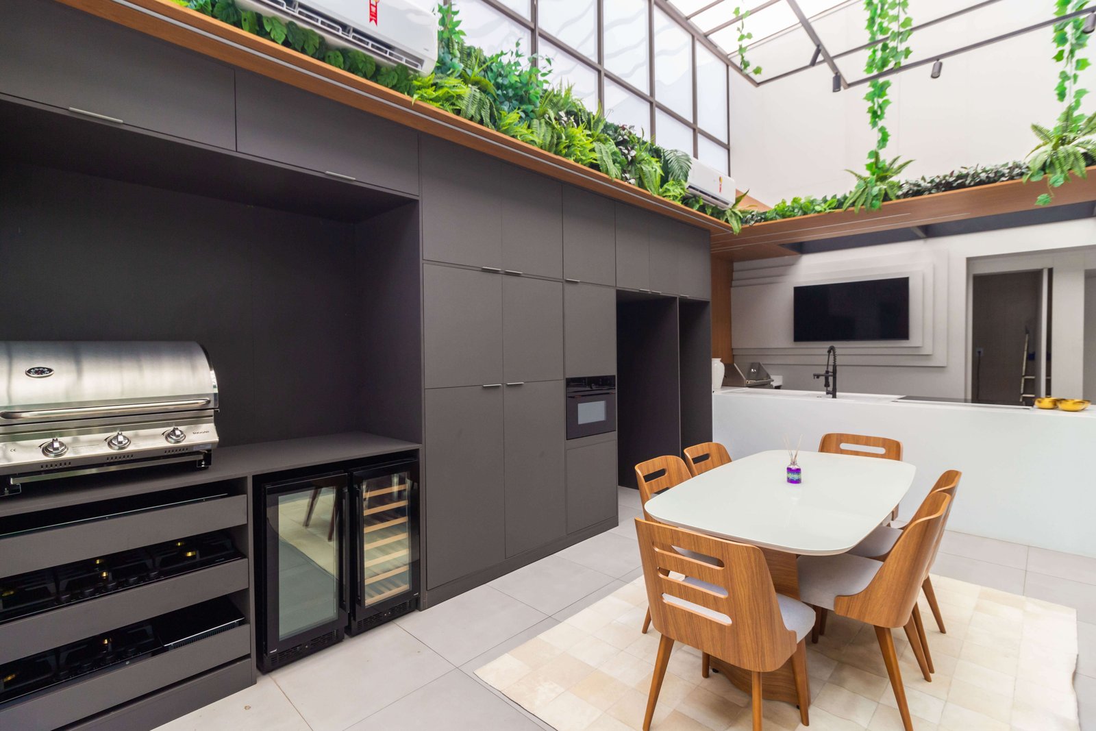 Cozinha gourmet integrada com armários escuros e bancada branca, equipada com adega climatizada e mesa de jantar, em ambiente com teto de vidro e plantas.