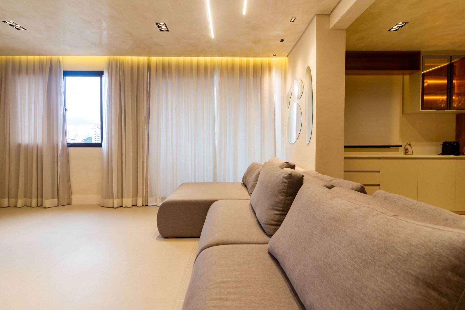 Sala de estar com sofá em L modular, cortinas fluidas e iluminação indireta no teto, criando um ambiente acolhedor e moderno.