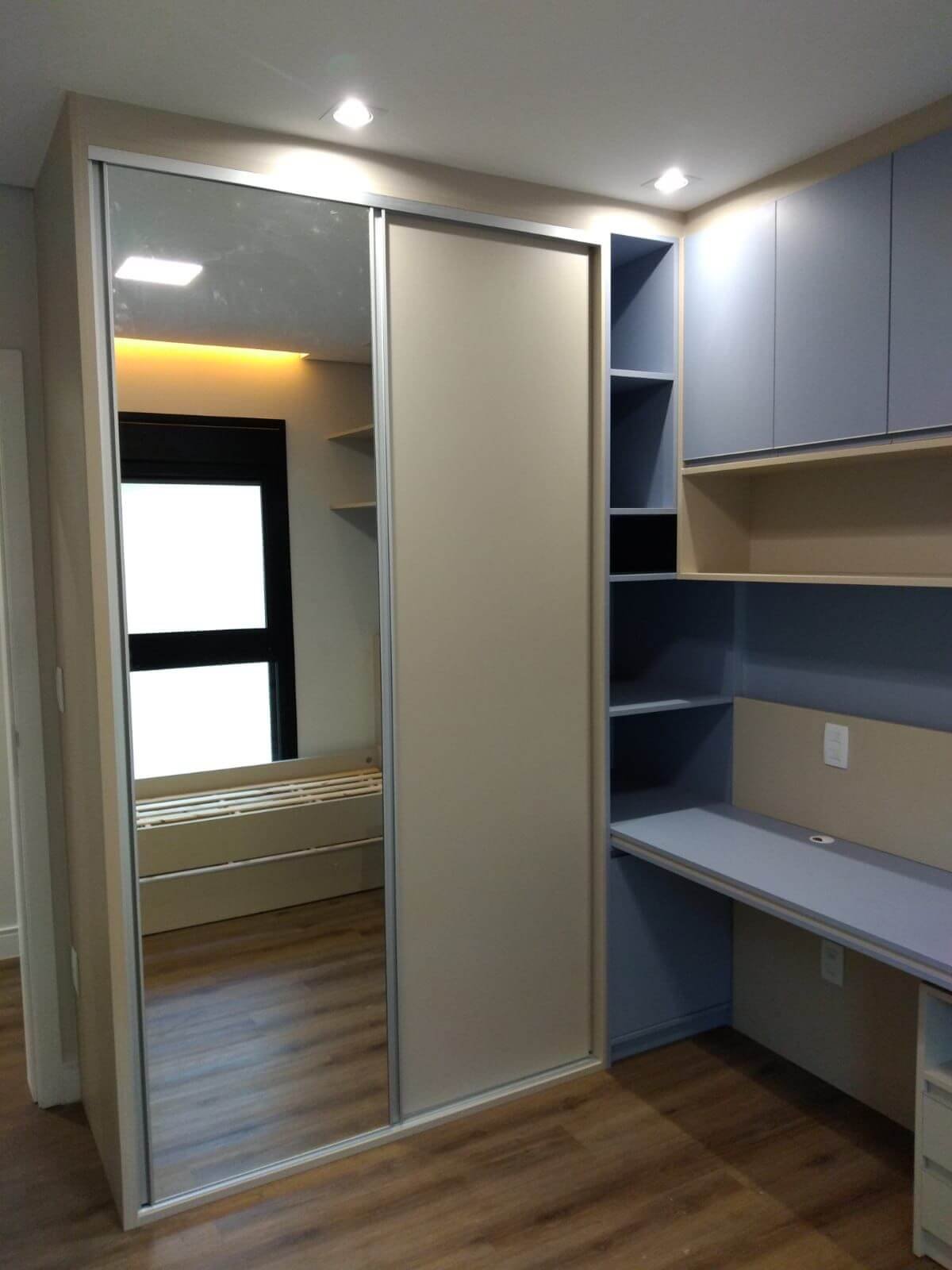 Um quarto com guarda-roupa de portas de correr (uma delas espelhada) e uma estante/escrivaninha planejada em tom azul-acinzentado ao lado. O piso é de madeira.
