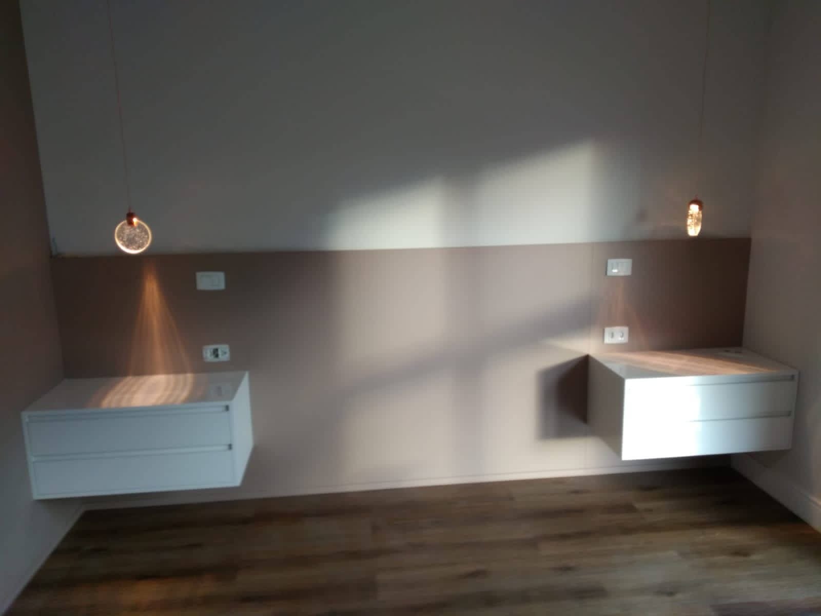 Um quarto vazio focado na parede da cabeceira, que possui um painel bege cobrindo a metade inferior da parede, duas mesas de cabeceira flutuantes brancas e arandelas de luz acesas em cada lado.