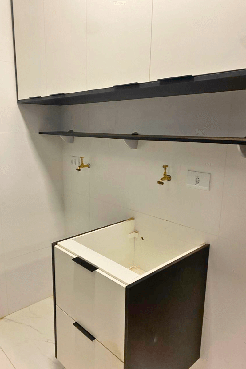 Lavanderia em fase de instalação com tanque branco embutido em gabinete preto e branco, prateleira superior preta e torneiras douradas na parede.