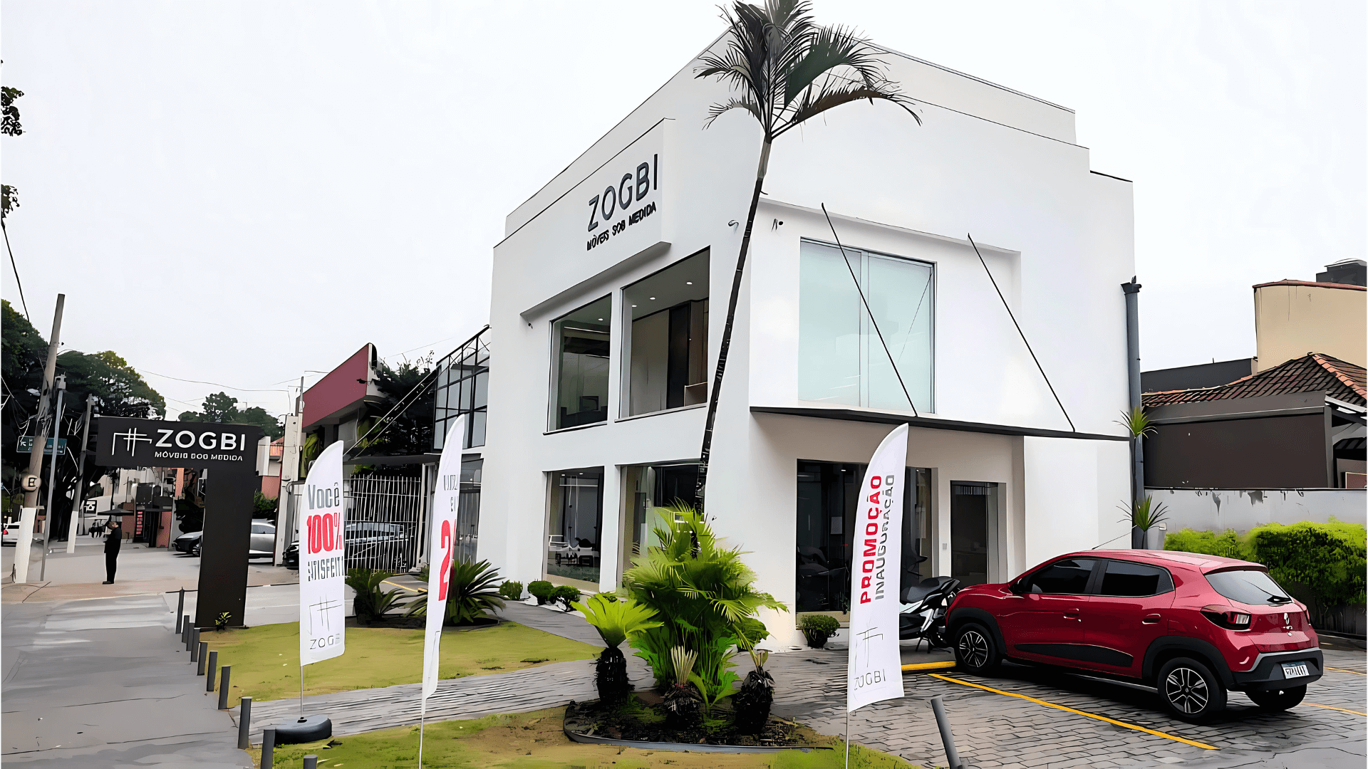 Fachada moderna da loja Zogbi Móveis Sob Medida localizada no Parque Ibirapuera na capital paulista em um dia nublado, com um carro vermelho estacionado na frente e bandeiras promocionais ao longo do caminho.
